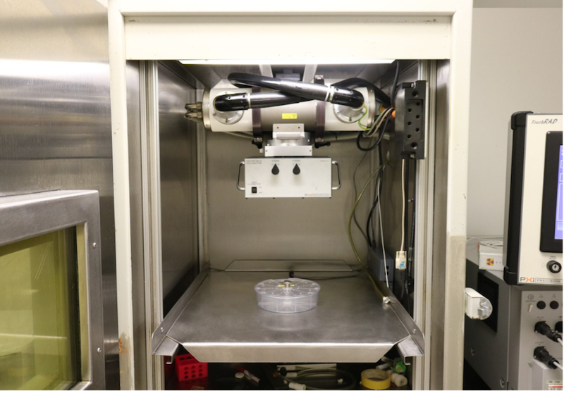 Laboratory setup featuring an X-RAD irradiator with a see-through dish on a metal shelf. A portion of another machine with a display screen and control knobs is visible on the right.